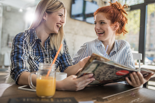 Two female friends reading something funny in a magazine and laughing while spending a day together in cafe.