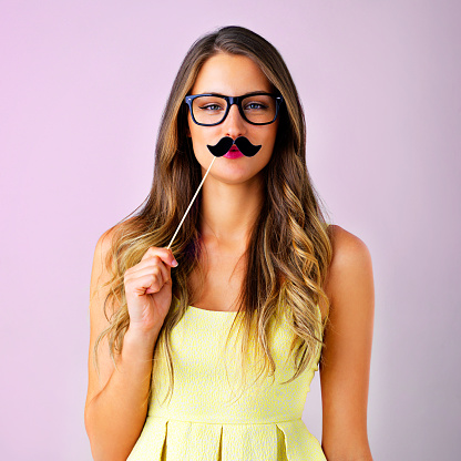 Studio shot of a young woman holding a mustache prop to her face against a pink background