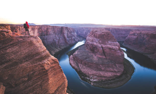 A male hiker is standing on steep cliffs enjoying the beautiful view of Colorado river flowing at famous Horseshoe Bend overlook in beautiful post sunset twilight on a summer evening, Arizona, USA
