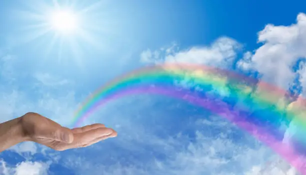 Male hand palm up with a rainbow appearing to end in his palm on a wide blue sky background with a bright sunburst overhead