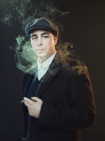 Portrait of stylish gangster wearing suit standing and smoking cigarette. Real man in peaky blinders style suit against black background. Photography from a DSLR camera. Sharp focus on eyes.