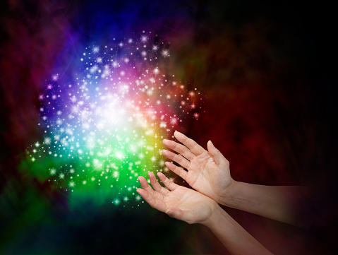 Female hands reaching towards a cloud of rainbow colored sparkles on a dark background with copy space
