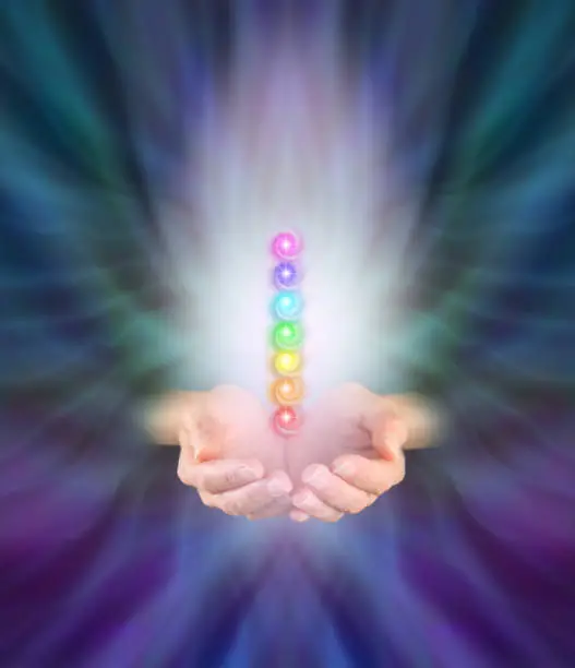 Healer's hands emerging from feather like multicolored energy formation background, cupped with seven chakra vortexes hovering above in a misty light with plenty of copy space