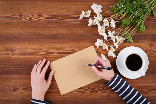 Top view woman hands writing on the blank paper sheet with blank copy space for text, white flowers and cup of coffee on wooden background