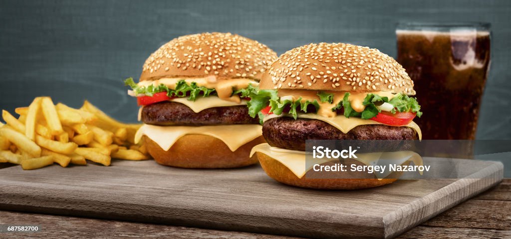 Two craft beef burgers on wooden table on blue background Two craft beef burgers on wooden table isolated on blue background. A glass with a drink stands in the background with a french fries Burger Stock Photo
