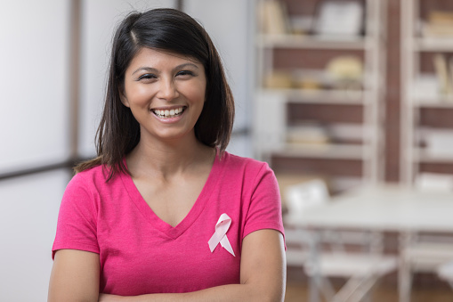 Cheerful mid adult Asian breast cancer survivor wears a breast cancer awareness ribbon on her t-shirt. She is smiling at the camera.