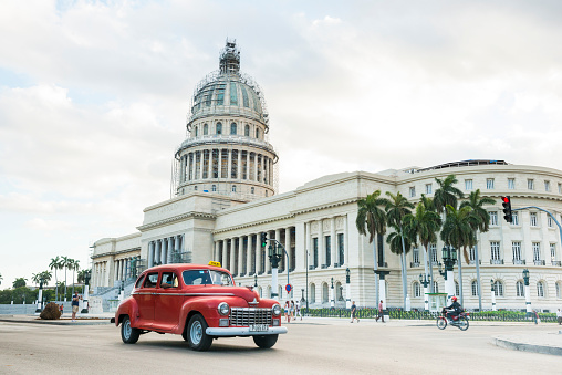 This is a horizontal, color photograph of a red Dodge classic car taxi driving on the streets of travel destination Havana, Cuba. People are outside on the street in front of the landmark El Capitolio Building, which has scaffolding around the dome. Photographed with a Nikon D800 DSLR camera. CreativeContentBrief 696189239