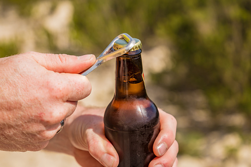 A man is standing in nature and is opening a bottle of beer with a bottle opener.