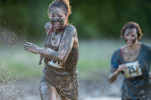 Beautiful young woman with blonde hair covered in mud running through splashing water wearing a race bib and muddy casual clothing during a mud run outdoors in the summer.