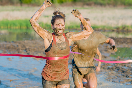 Beautiful blonde young woman leaping through the finish line with her arms up and smiling in celebration while competing in a mud run for charity.