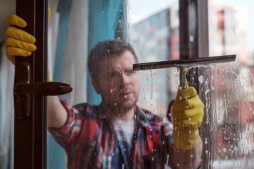 A man is washing a window on the balcony. He holds a scraper in his hands and carefully rubs the glass