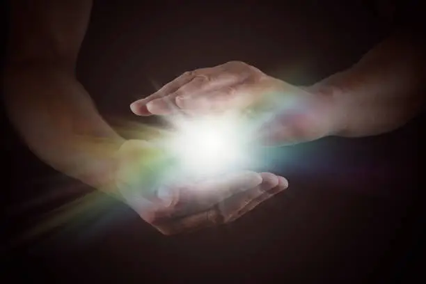 Male hands emerging from darkness, cupped, with rainbow colored bright white star burst of light between