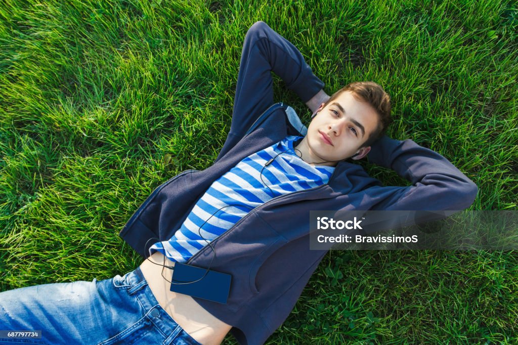 Young man listening music from smart phone on the grass in the park. Teenage man in park in spring, listening music with headphones, smart phone on grass. Rest and dream. Grass Stock Photo