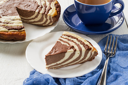 Delicious homemade chocolate cake with cottage cheese Zebra (Marble cake). Tasty breakfast. Piece of cake on blue plate, blue cup and blue gauze textiles on white wooden table. Selective focus