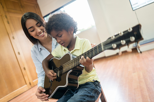 Boy at the school playing the guitar in music class and teacher supervising him - education concepts