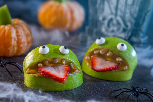 Healthy Halloween candy alternative fruit.  Apple monsters made of peanut butter, sunflower seeds and candy eye balls with spiders, spider webs, and tangerine pumpkins with celery stem on haunted halloween night.