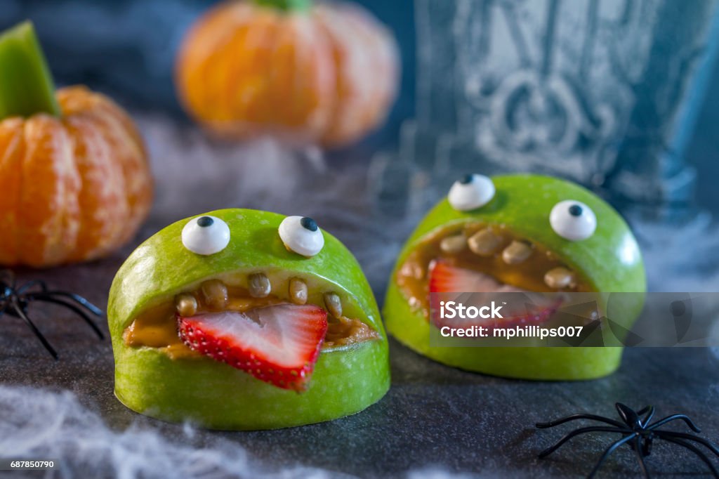 Healthy Halloween Apple Monsters Fruit Kids Treat Healthy Halloween candy alternative fruit.  Apple monsters made of peanut butter, sunflower seeds and candy eye balls with spiders, spider webs, and tangerine pumpkins with celery stem on haunted halloween night. Halloween Stock Photo