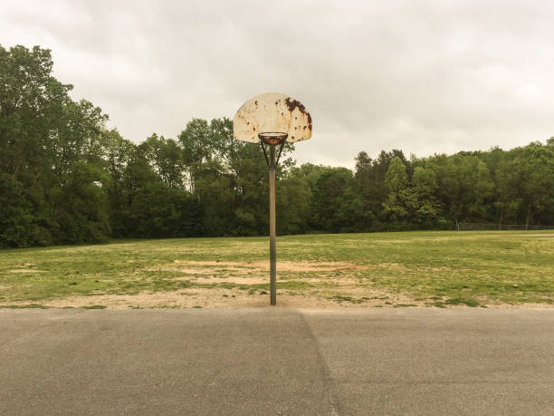 On a very cloudy morning an outdoor public basketball court lays unused, has been for some time. Tall weeds grow up the fences. The hoop and backboard and posts are rusty.