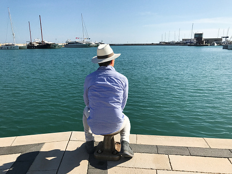 Senior Man in White Hat in Mediterrean Port (Back View). He is sitting, contemplating, and enjoying the view of the sea and boats. Enjoying his retirement. Copy space available.