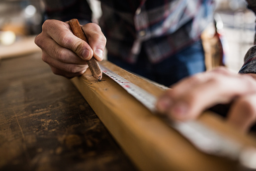 Close up of male worker using pen while drawing measurements on a wood plank in a carpentry workshop.