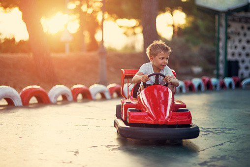 Little boy aged 6 riding a bumper car in funfair amusement park. The boy is holding the steering wheel with confidence and his eyes are focused on the road. Sunny summer day is ending.
