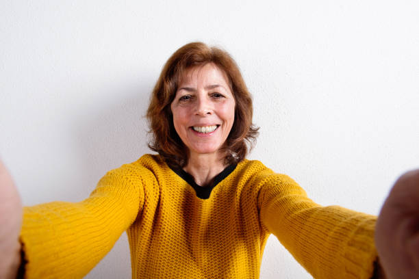 Senior woman in yellow sweater, taking selfie, studio shot.