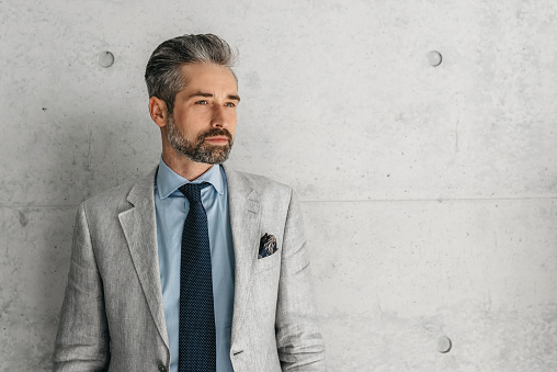 handsome bearded businessman in gray business Sakko standing  in front of concrete wall