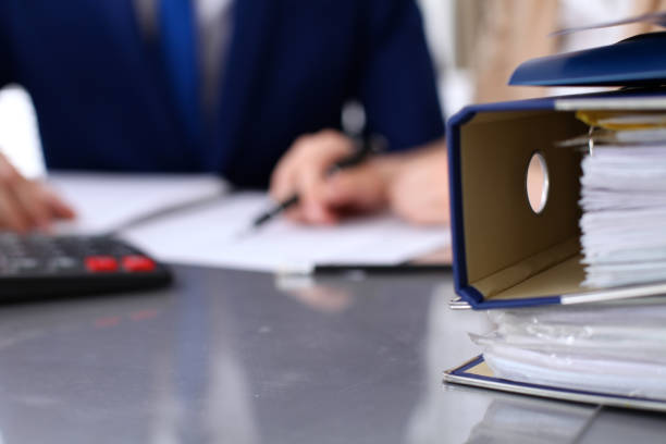 istock Binders with papers are waiting to be processed with businessman and secretary back in blur. Internal Revenue Service inspector checking financial document. 688002530