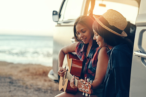 Shot of a young woman playing a guitar on a road trip with her friend