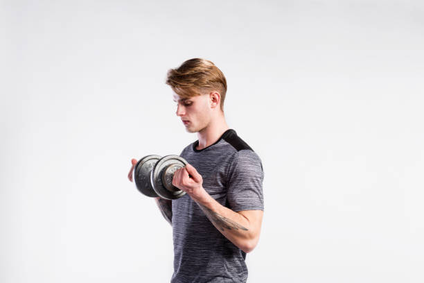 Fitness man holding dumbbell, working out, studio shot. Handsome hipster fitness man in gray t-shirt working out with dumbbell. Studio shot on gray background. forearm tattoos men stock pictures, royalty-free photos & images