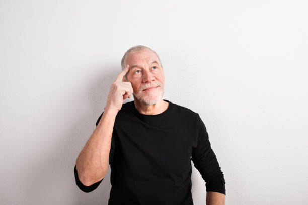 Thoughtful handsome senior man in black sweater, finger on forehead. Studio shot against white wall.