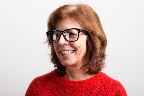 Beautiful senior woman in in black eyeglasses and red sweater, smiling. Studio shot against white wall.