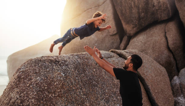 Father and son enjoying summer holidays at the beach Shot of cute little boy leaping into his fathers arms from a big rock. Father and son enjoying summer holidays at the beach. catching stock pictures, royalty-free photos & images