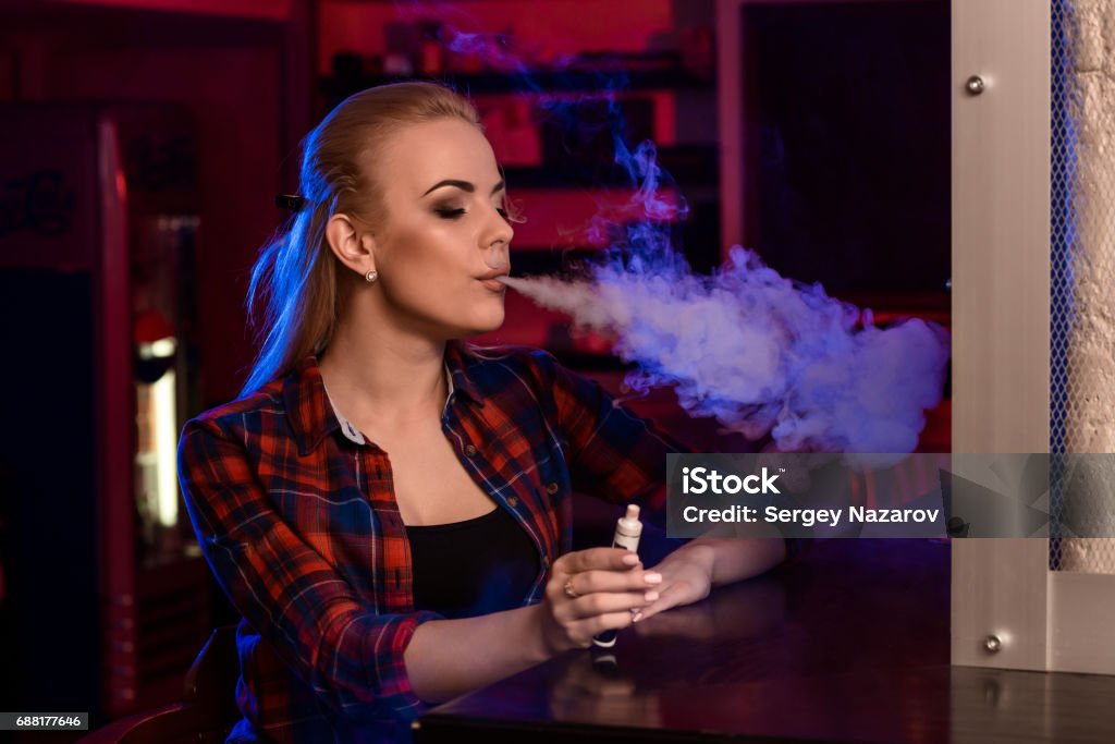 Young pretty woman in a shirt in a cage smoke an electronic cigarette at the vape bar Young pretty woman in a shirt in a cage smoke an electronic cigarette at the vape bar. Vape shop. Addiction Stock Photo