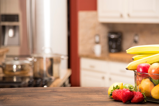 Background of a defocused home kitchen with a wooden table in foreground for object placement or copyspace.  A bowl of fruit at side.  No people.