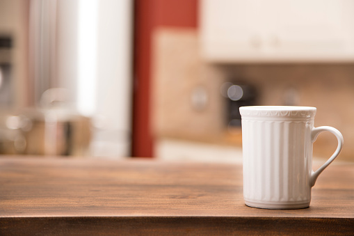 Background of a defocused home kitchen with a wooden table in foreground for object placement or copyspace.  A white coffee mug at side.  No people.