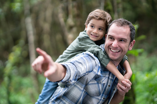 Portrait of a happy father carrying his son outdoors on a piggyback ride and pointing away - lifestyle concepts