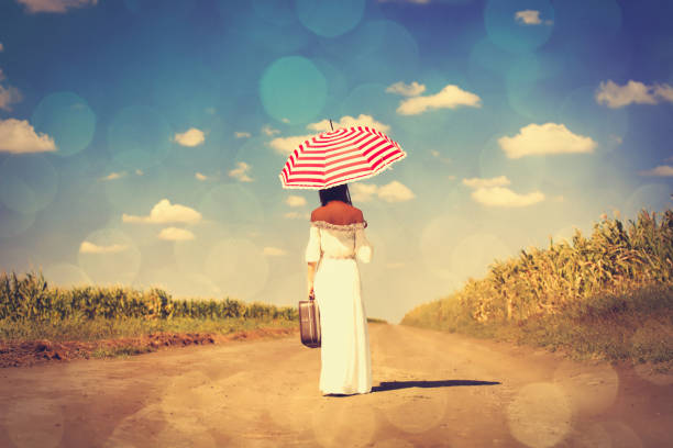 photo of beautiful young woman with umbrella and suitcase on the wonderful road on sky and field background