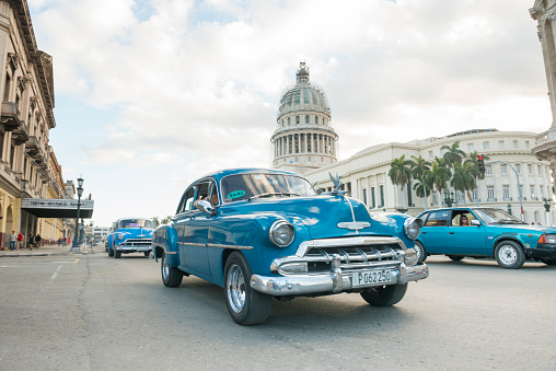 This is a horizontal, color photograph of an blue Chevrolet classic car from the 1950s driving as a taxi on the streets of travel destination Havana, Cuba. People are visible in the cars in front of the landmark El Capitolio Building, which has scaffolding around the dome. Photographed with a Nikon D800 DSLR camera. CreativeContentBrief 696189239