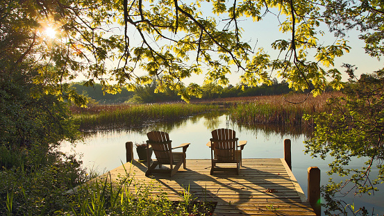 Relaxing setting with two adirondack chairs on a pier over looking a calm and tranquil lake at sunrise with reflections on a Spring day