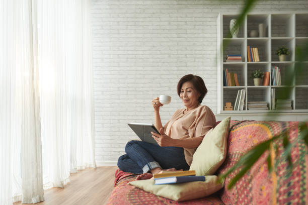 Resting at home Vietnamese senior woman drinking tea and reading book on tablet computer south east asian ethnicity stock pictures, royalty-free photos & images