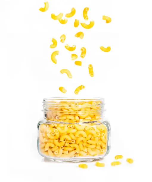 raw macaroni pasta in clear glass jar with pieces falling down into the jar and some on the ground, on isolated white background