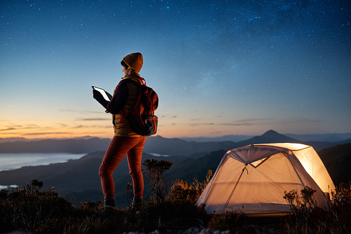 Shot of a young hiker using a tablet while standing in her campsite at the top of a mountain