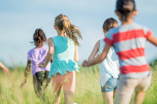 A multi-ethnic group of elementary school boys and girls are outdoors. They are in a field in a rural environment. They are wearing casual clothes. It is springtime and sunny. The children are running in the field. They are following a girl of African descent.