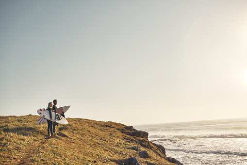 Shot of a young couple walking up a cliff at the beach with their surfboards