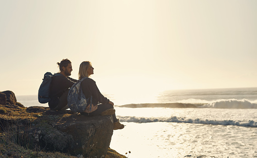 Shot of a young couple admiring the view while sitting on a cliff at the beach