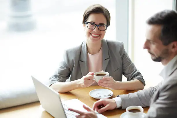 Happy young businesswoman with cup of tea looking at camera with co-worker sitting near by