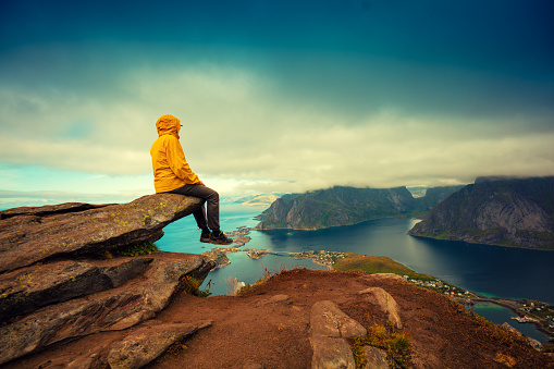 Panoramic aerial view of fjord and fishing village. Man tourist sitting on a cliff of rock. Beautiful mountain landscape. Nature Norway, Lofoten islands.