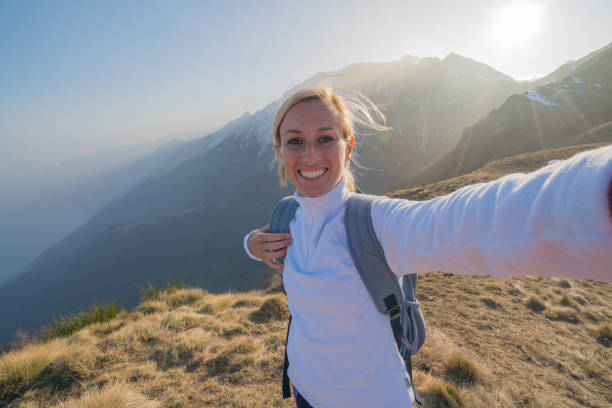 Young woman hiking reaches mountain top and takes a selfie with the mountain landscape on the background. Shot in Switzerland, springtime.