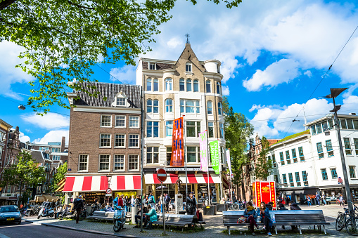Amsterdam, Netherlands - May 28, 2015: Street with the house in Amsterdam on a sunny day. People are sitting on benches in a small park in Amsterdam. Horizontal photo. Blue sky, green blooming trees.